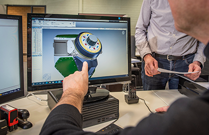 Person in a black sweater points at a 3D CAD model of a mechanical part on a monitor while colleagues stand nearby in an office.
