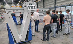 Eine Gruppe von Arbeitern und ein Ausbilder inspizieren eine große graue Industriemaschine in einer Fabrikumgebung. Im Vordergrund steht ein blauer Wagen mit großen Platten.