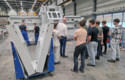 Group of workers and an instructor inspecting a large gray industrial machine in a factory setting, with a blue cart carrying large panels in the foreground.