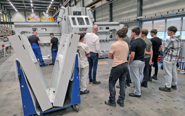 Eine Gruppe von Arbeitern und ein Ausbilder inspizieren eine große graue Industriemaschine in einer Fabrikumgebung. Im Vordergrund steht ein blauer Wagen mit großen Platten.